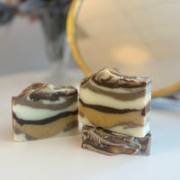 Three marbled artisanal soap bars with layers of white, dark brown, and tan, displayed on a white surface with a blurred mirror and flowers in the background.
